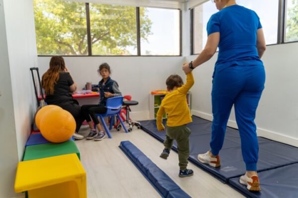 A nurse guiding a toddler walking on floor mats while other children work at an activity table at Little Giants PPEC