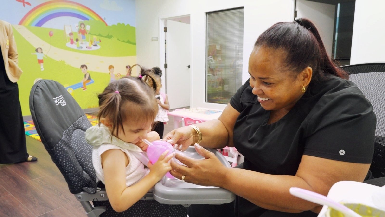 Smiling caregiver helping a toddler drink from a sippy cup in a high chair at Little Giants PPEC in Tampa, FL