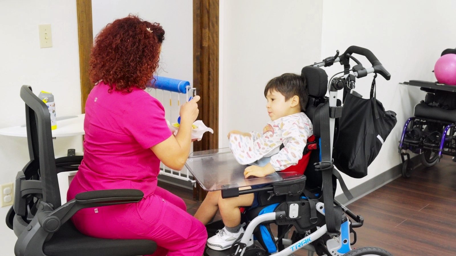 Caregiver in pink scrubs doing a tabletop activity with a boy in an adaptive chair at Little Giants PPEC in Tampa, FL