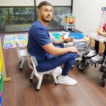 Staff member sitting with a school-age boy in a wheelchair at a play table in the playroom at Little Giants PPEC in Tampa, FL