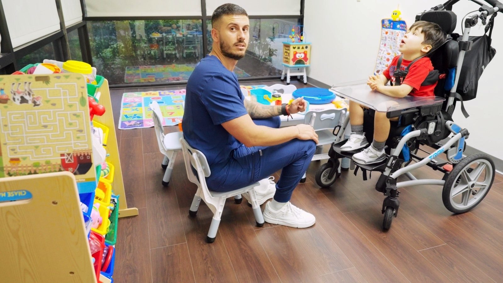 Staff member sitting with a school-age boy in a wheelchair at a play table in the playroom at Little Giants PPEC in Tampa, FL